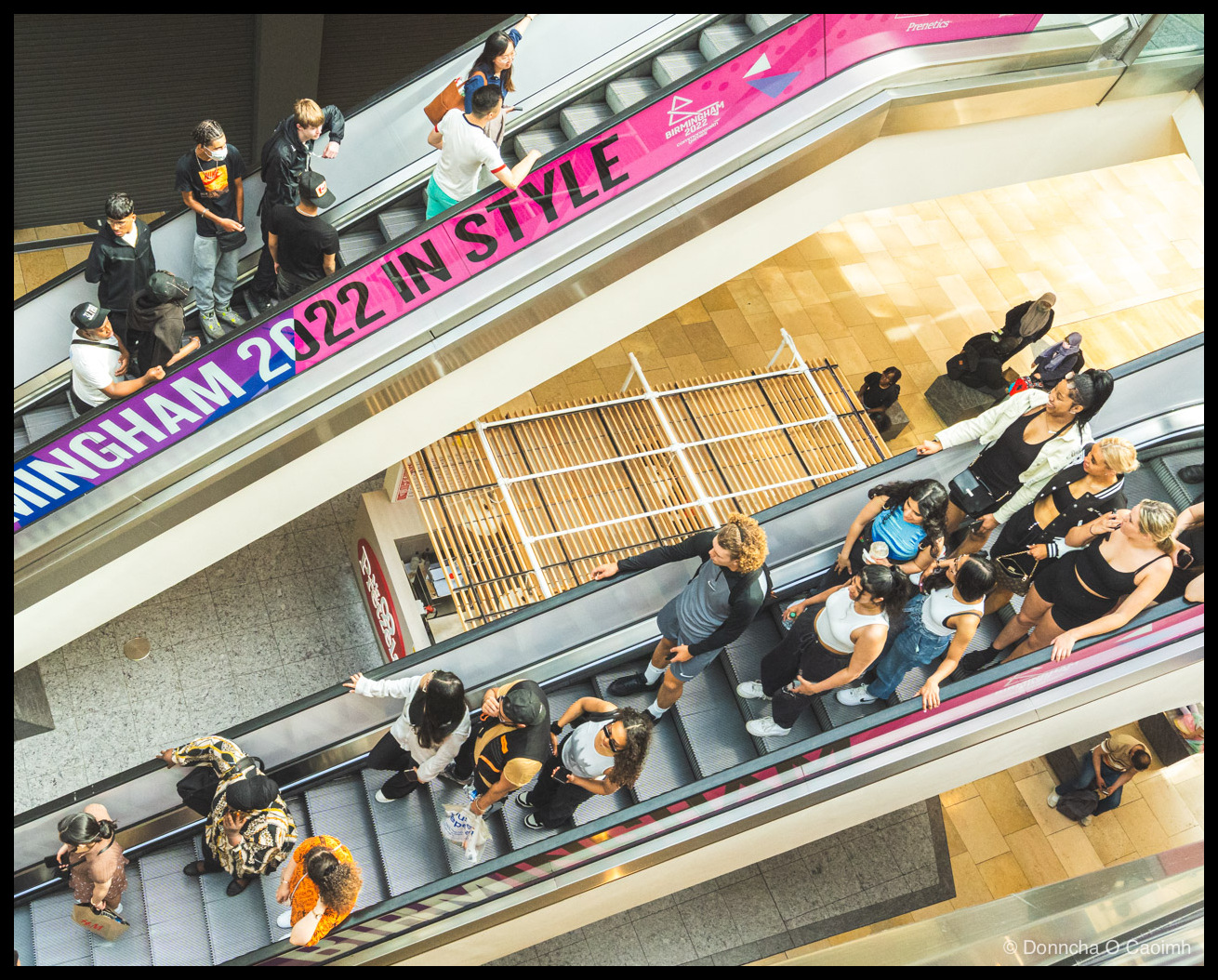 Photo shot from above of two escalators crisscrossing through a shopping centre, packed with people going up and down, the upper handrail carrying a purple banner reading "...MINGHAM 2022 IN STYLE" and "BIRMINGHAM 2022" with a small "Prenetics" logo.