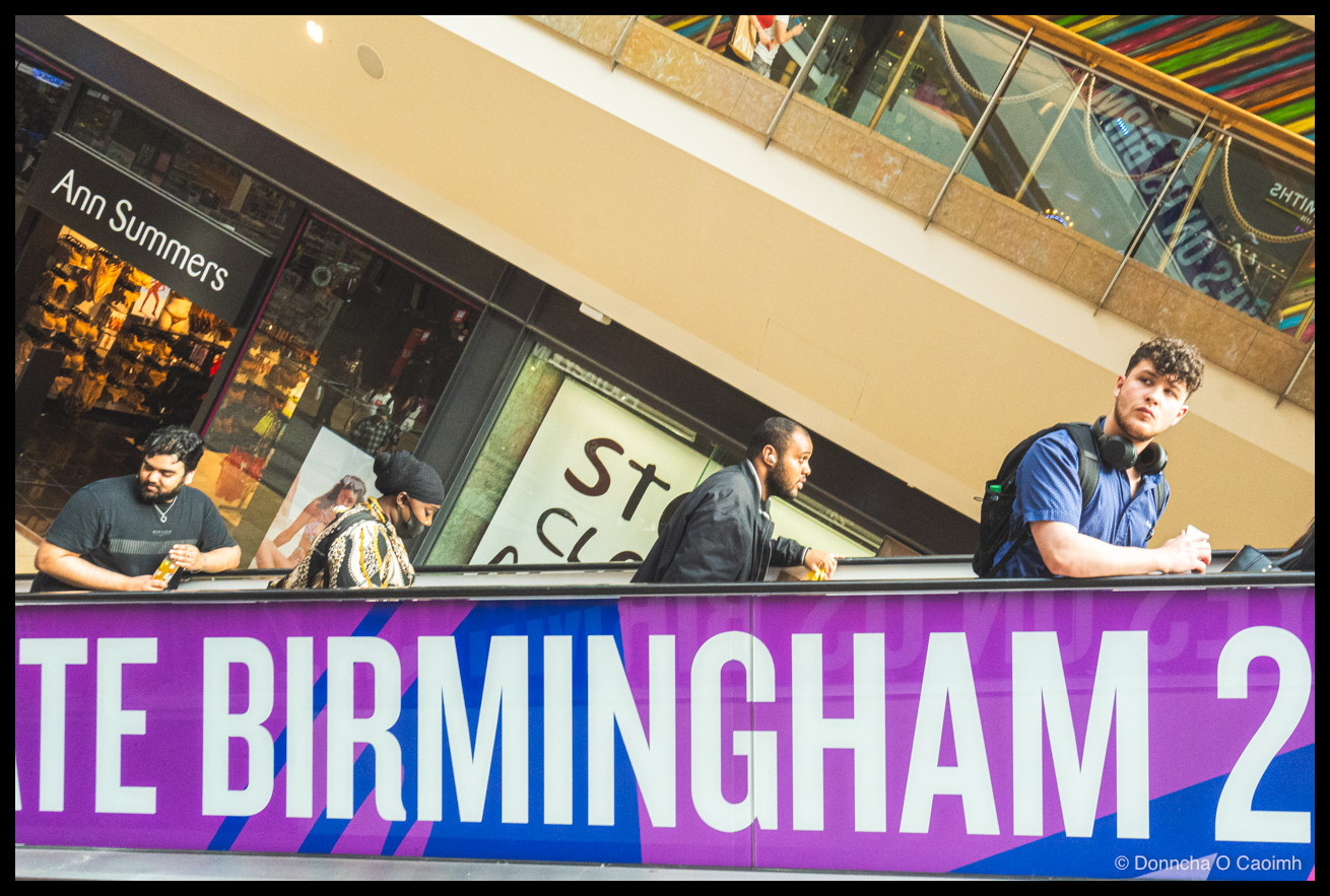 Four people leaning on an escalator, the rail edged with a purple "...TE BIRMINGHAM 2..." Commonwealth Games banner, an Ann Summers shopfront to the left and a young man in a blue shirt with headphones round his neck staring toward the camera.