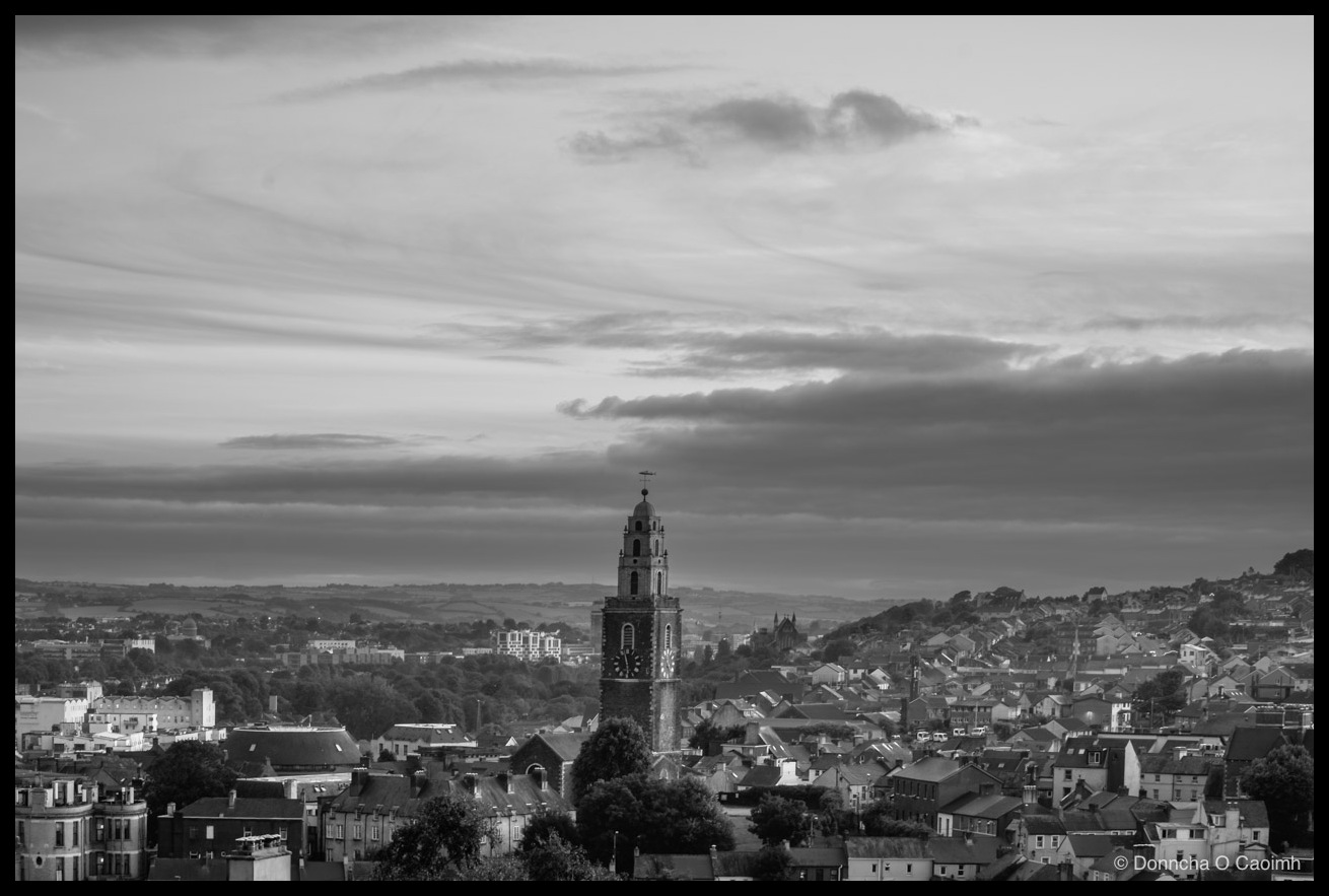 Black-and-white photo looking across the rooftops of Cork City's north side at dusk, with the tower of St Anne's Church, Shandon, rising above the houses, its clock face and stepped belfry catching the light beneath a broad sky of streaked cloud, distant hills along the horizon.