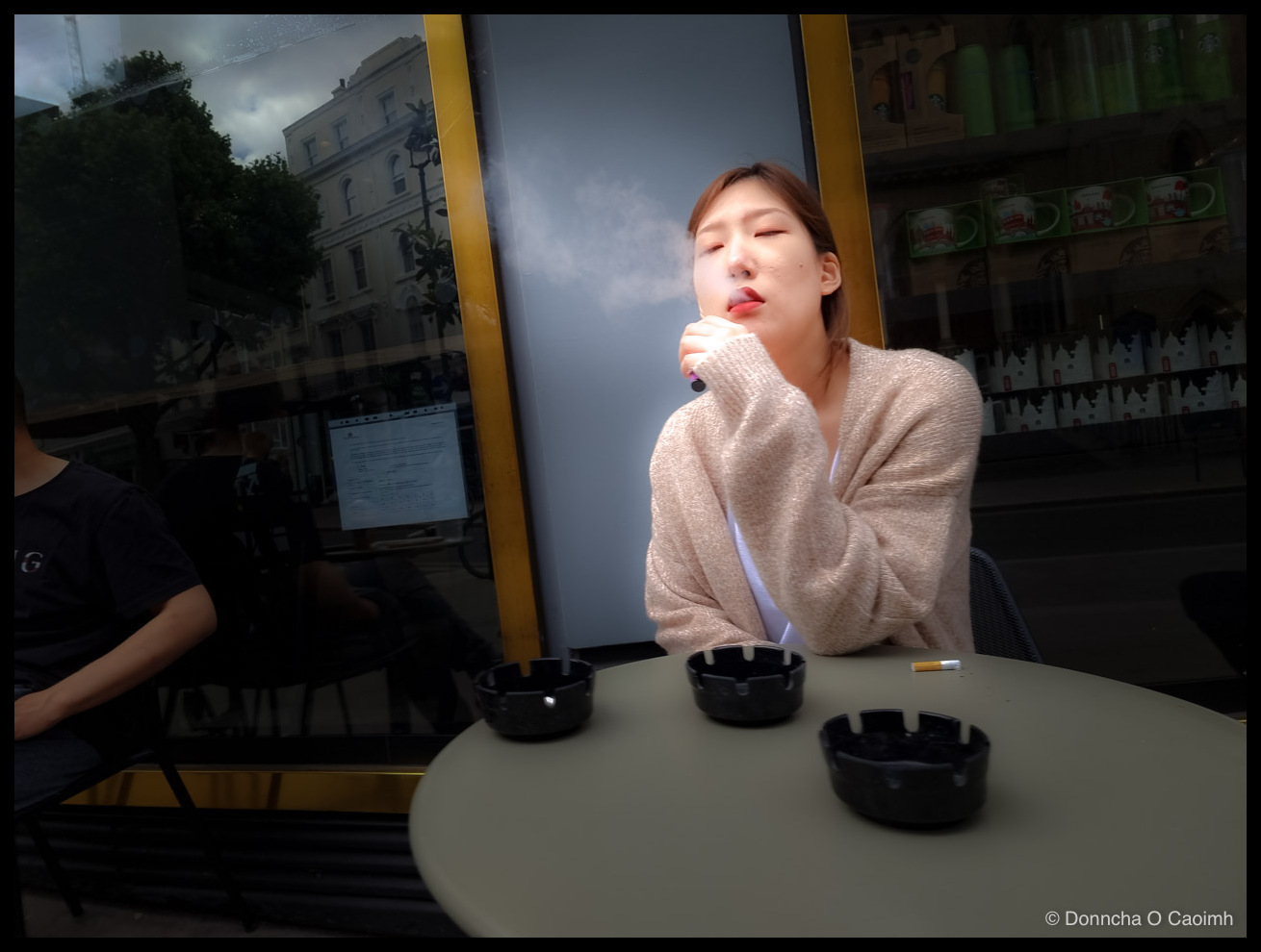 A young woman seated at an outdoor café table in London, eyes closed, exhaling a cloud of vapour from a vape held to her lips. She wears a fawn knitted cardigan over a white top. Three black ashtrays and a discarded cigarette filter sit on the round cream table. A man in a black T-shirt sits at the left, and a large café window behind reflects a cloudy street of period buildings while showing the interior shelves of a Starbucks store.