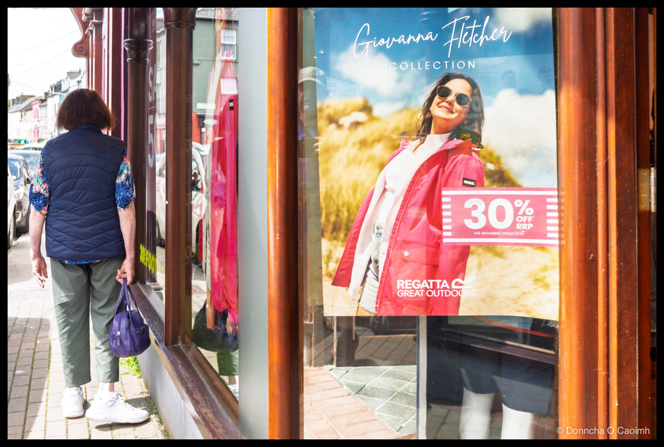 A woman with short brown hair seen from behind, wearing a navy gilet over a blue floral top, olive trousers and white trainers, carrying a purple handbag as she gazes into a shop window beside a sunlit Regatta poster reading "Giovanna Fletcher COLLECTION", "30% OFF RRP THE GIOVANNA COLLECTION" and "REGATTA GREAT OUTDOORS".