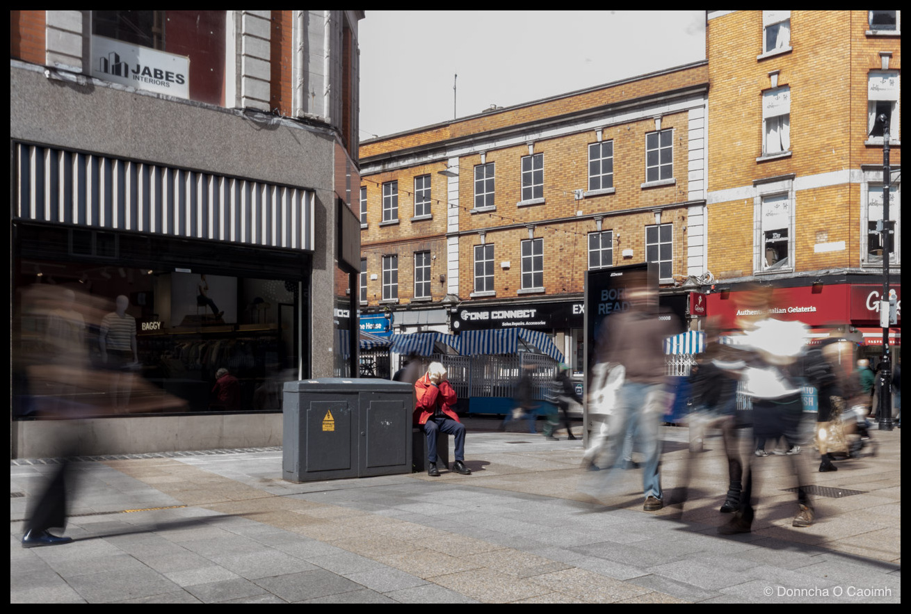 Colour street photo on Henry Street, Dublin: a man in a red jacket sits on a grey utility box with a yellow electrical hazard sticker, raising a hand to his face, while blurred shoppers walk past; shop signage reads "JABES INTERIORS", "BAGGY", "ne House", "FONE CONNECT Sales.Repairs.Accessories", "EX", "Authentic Italian Gelateria", "Gi", and a digital screen reads "BOR READ"