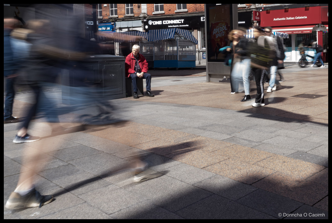 Colour street photo on Henry Street, Dublin, in strong sunlight: a man in a red jacket sits calmly on a black utility box with clasped hands as blurred pedestrians and long shadows cross the granite paving; signage reads "FONE CONNECT   Sales.Repairs.Accessories", "EX", "ne House", "Authentic Italian Gelateria", "Gino's", and an Aperol Spritz advertisement with "CREPES WAFFLES COFFEE"