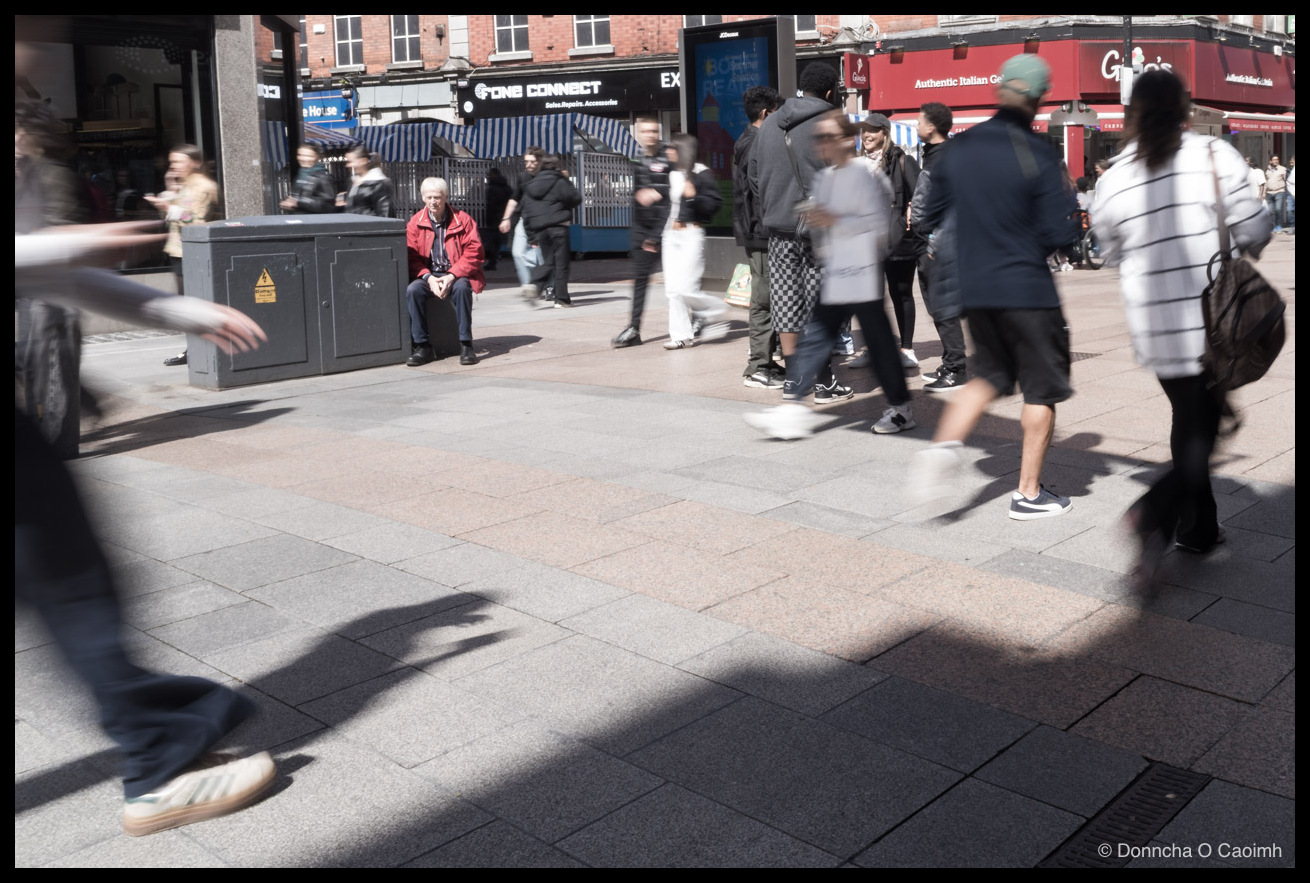 Colour street photo on Henry Street, Dublin: a man in a red jacket sits on a grey utility box surrounded by a busy crowd, some standing still and others blurred mid-stride across the sunlit pavement; signage reads "FONE CONNECT Sales.Repairs.Accessories", "EX", "ne House", "Authentic Italian Gelateria", "Gino's", and a digital screen showing "CREATE"