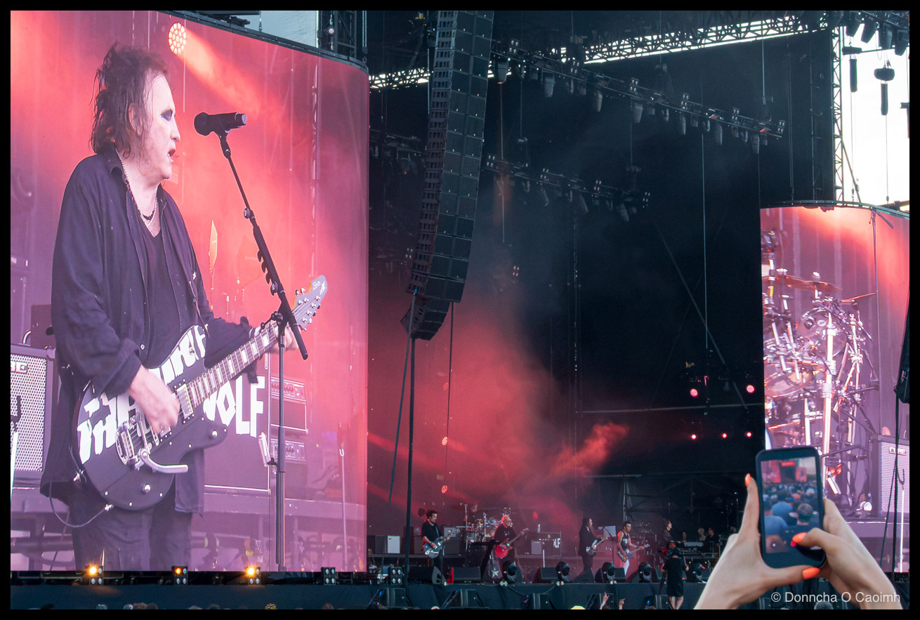 Robert Smith and The Cure on stage. Robert Smith on guitar on the big screen. Someone holds a photo up to take photos.