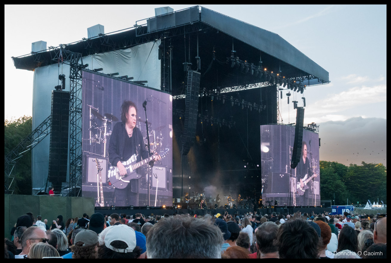 The stage, big screens. Robert Smith on the screens. Crowd in front.