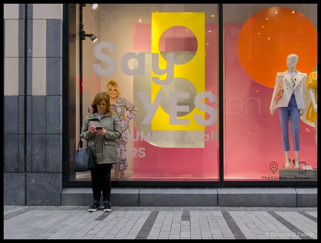 A woman in an olive green parka with a black tote bag standing on a paved street outside a shop window, head down and thumbing at a phone, while behind the glass a poster reads "Say YES" and "SUMMER of all ...ERS", with a mannequin in a white jacket and blue jeans to the right and small icons labelled "Click & Collect" and "Shop app".