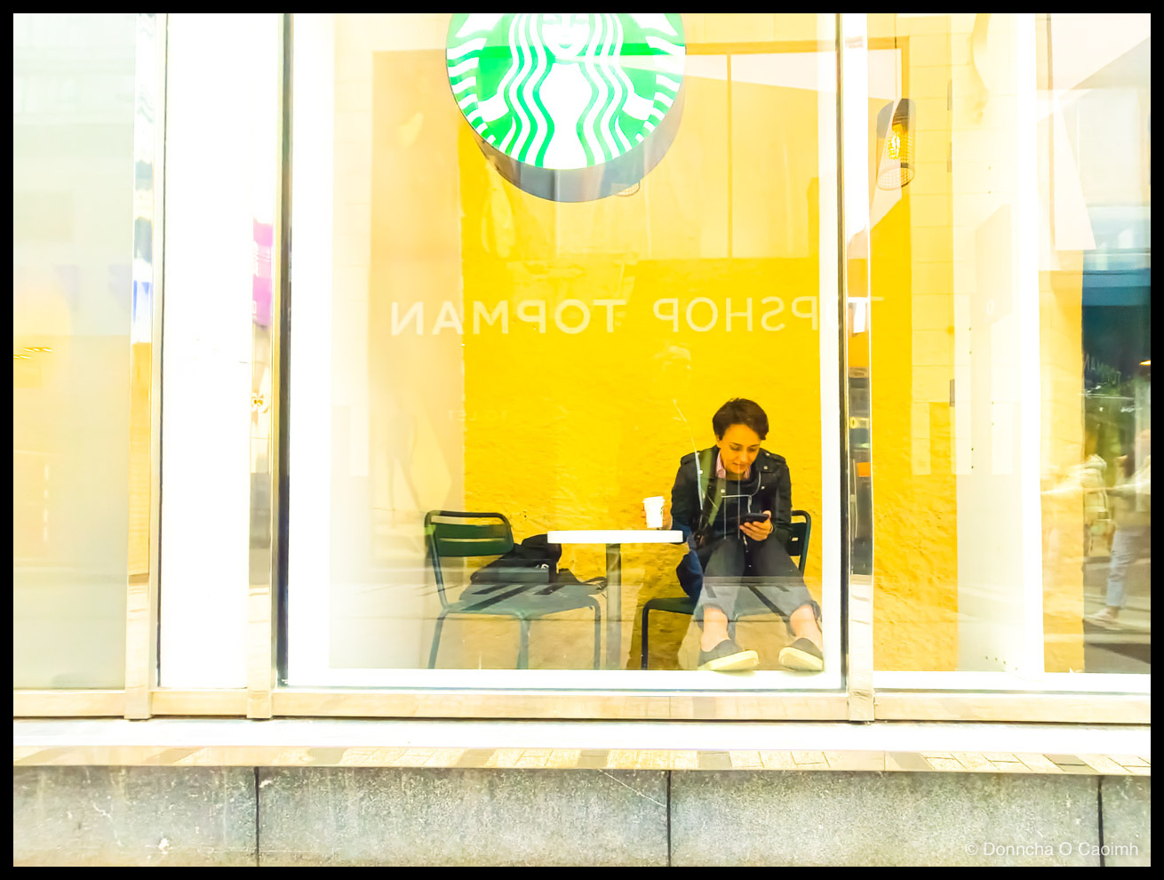 Photo taken through a bright window with a green Starbucks siren logo above it, showing a woman in a dark jacket sitting on a green metal chair against a yellow wall, earphones in, holding a paper cup in one hand and a phone in the other, with reversed lettering reading "TOPSHOP TOPMAN" on the glass.