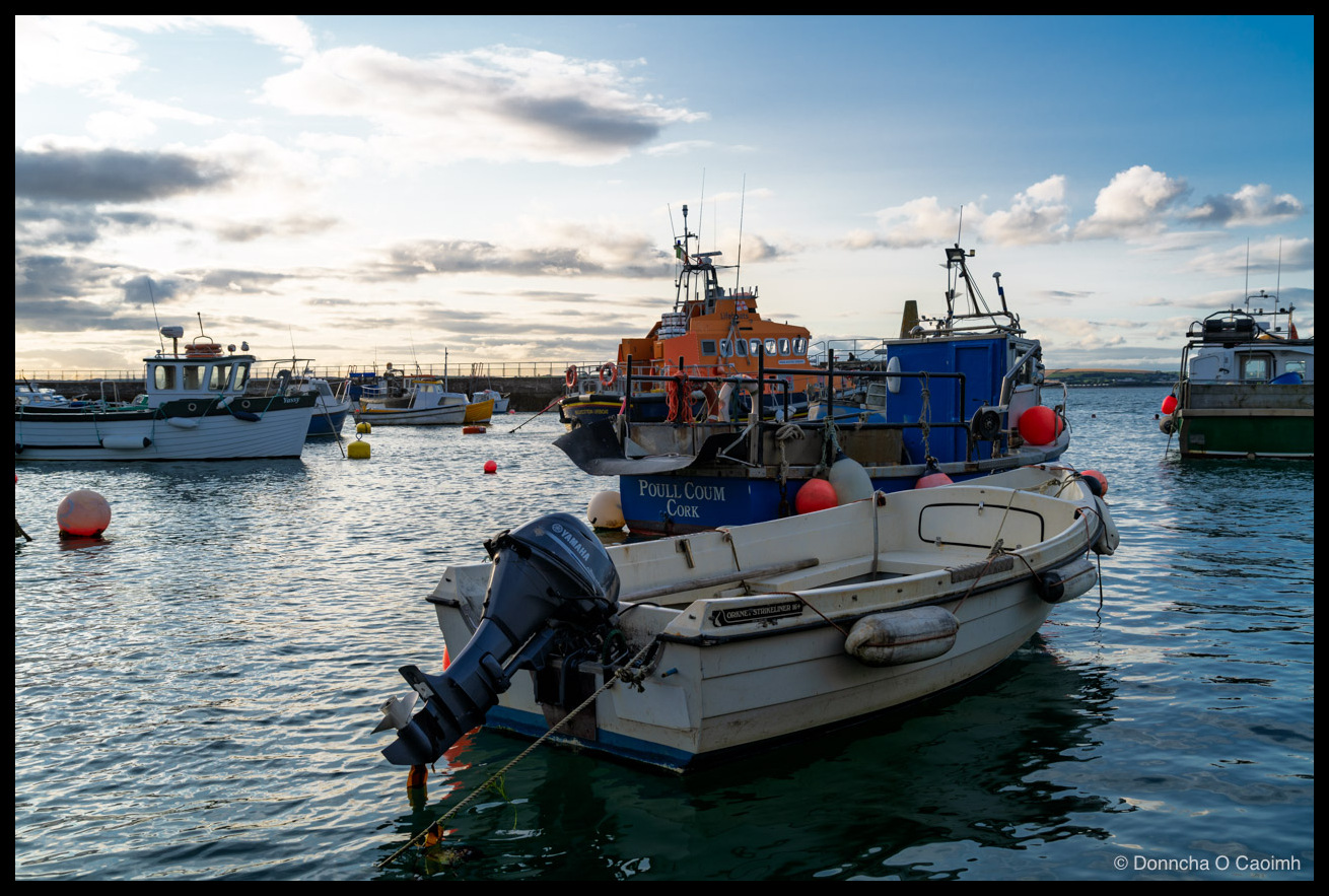 A harbour at dusk with a white dinghy and its Yamaha outboard motor in the foreground, a blue fishing boat marked "POULL COUM CORK" behind it, and an orange RNLI lifeboat and other small craft moored among red buoys under a soft evening sky.