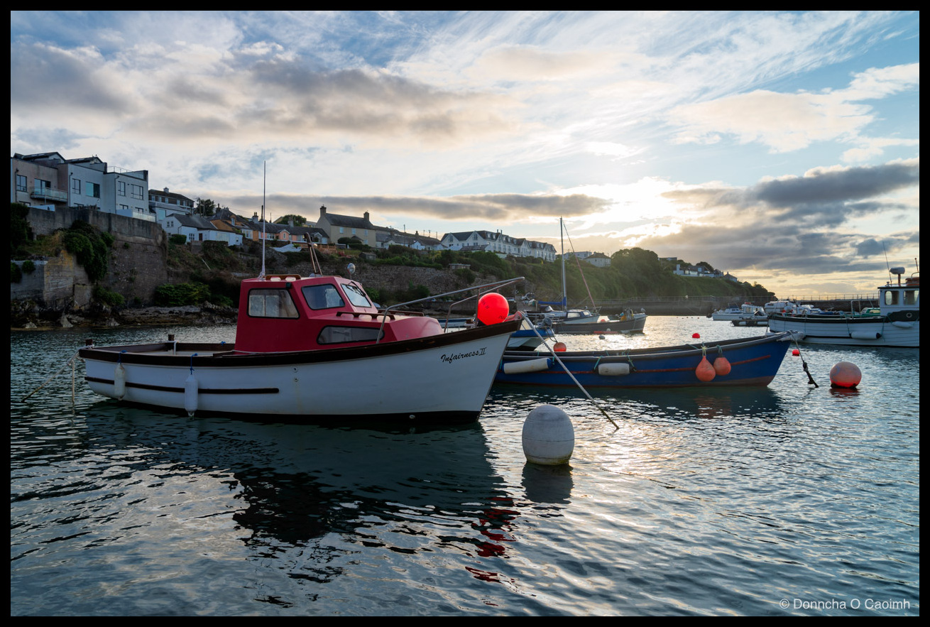 A red-and-white fishing boat named "Infairness II" moored in a calm harbour at sunset, with a hillside of houses to the left and small boats and buoys scattered across the water beneath a broken, glowing sky.