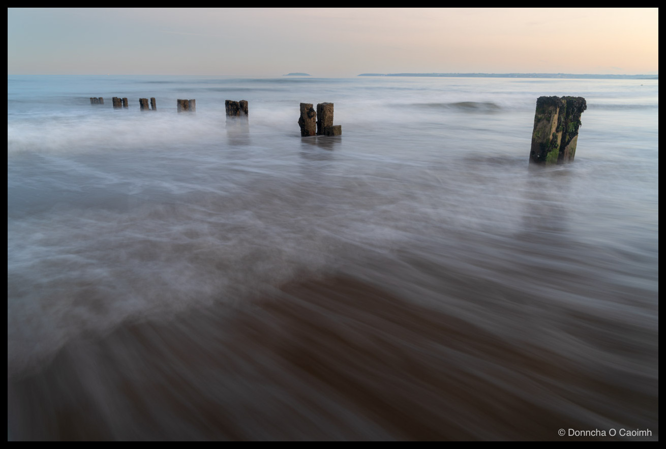 A long-exposure photograph of a line of weathered wooden groyne posts receding into a calm sea at Youghal beach; incoming water blurs into soft streaks over brown sand, with the nearest posts capped in green weed and a pale blue sky above.