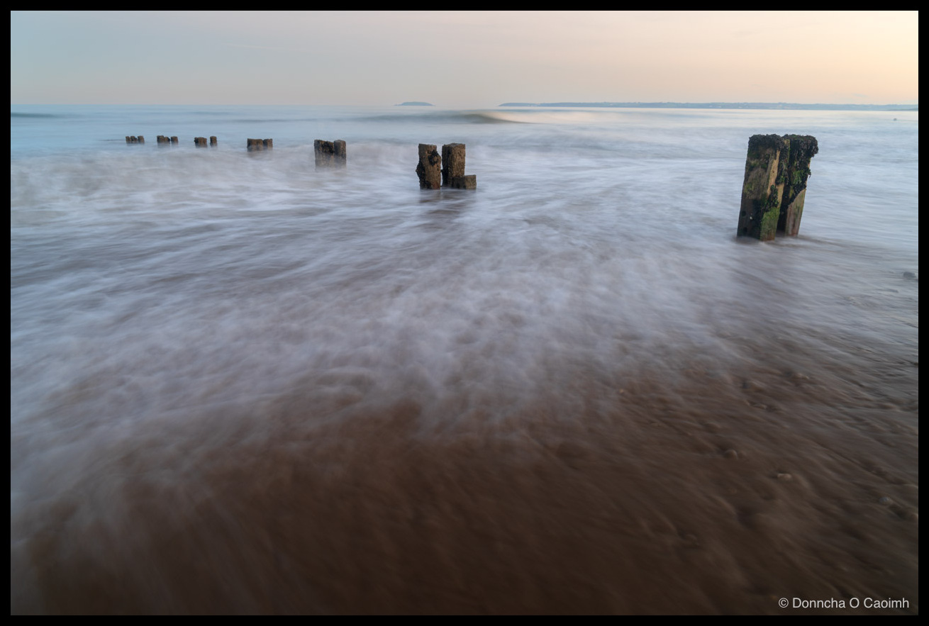 A long-exposure photograph of a line of weathered wooden groyne posts receding into a calm sea at Youghal beach; incoming water blurs into soft streaks over brown sand, with the nearest posts capped in green weed and a pale blue sky above.