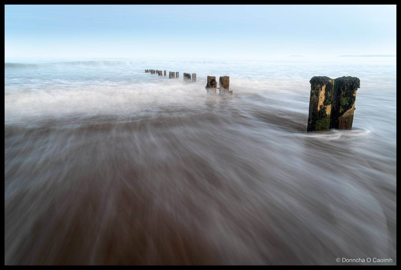 A long-exposure photograph of a line of weathered wooden groyne posts receding into a calm sea at Youghal beach; incoming water blurs into soft streaks over brown sand, with the nearest posts capped in green weed and a pale blue sky above.