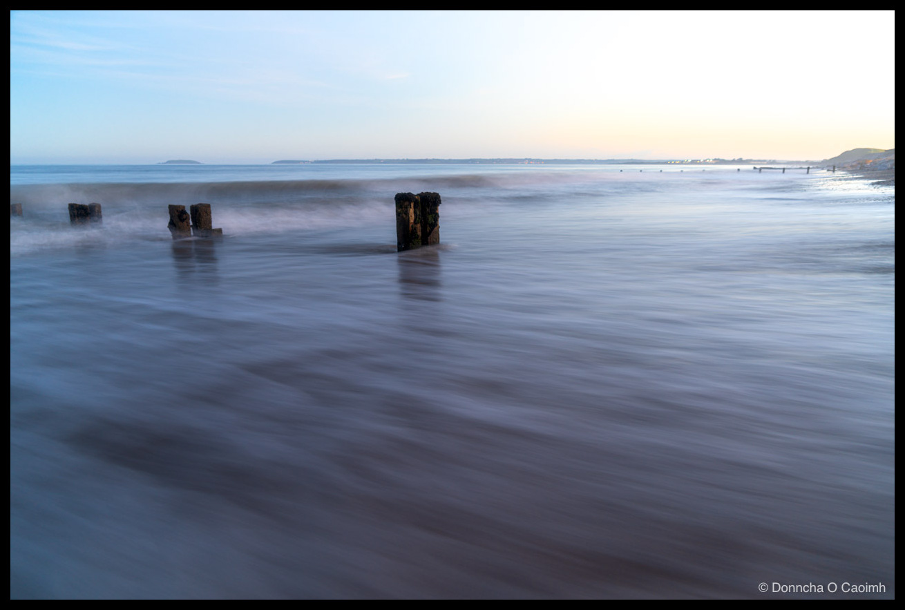 A long-exposure photograph of a line of weathered wooden groyne posts receding into a calm sea at Youghal beach; incoming water blurs into soft streaks over brown sand, with the nearest posts capped in green weed and a pale blue sky above.