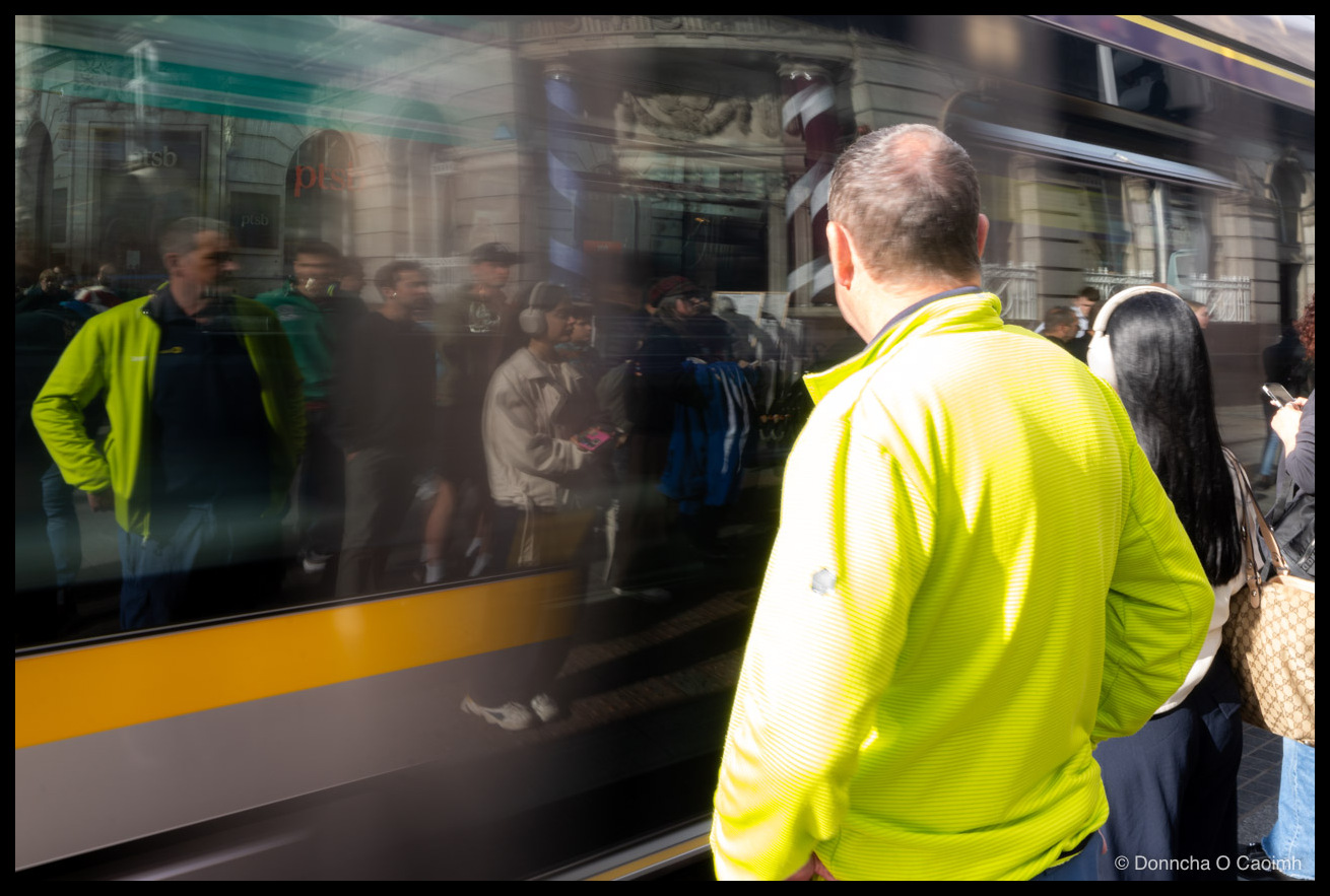 A Luas tram pulling away from O'Connell Street in Dublin, its side windows mirroring a blurred crowd of pedestrians against a grand stone building; a red-and-white barber's pole and a "ptsb" bank sign appear in the reflection, while a man in a lime hi-vis top and a woman with long dark hair stand in the foreground.