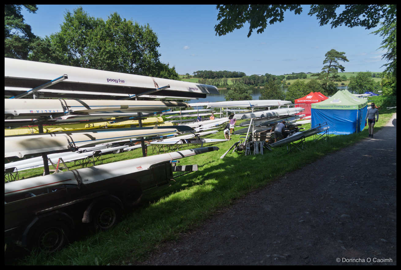 A long line of boat trailers stacked four high with white racing shells beside a reservoir, the nearest hulls marked "mj flood" and "ITALIA", with crews working on riggers in the middle distance and red, green and blue club gazebos marked "BANN ROWING CLUB" and "Commercial Rowing Club" pitched along the gravel path on the right.