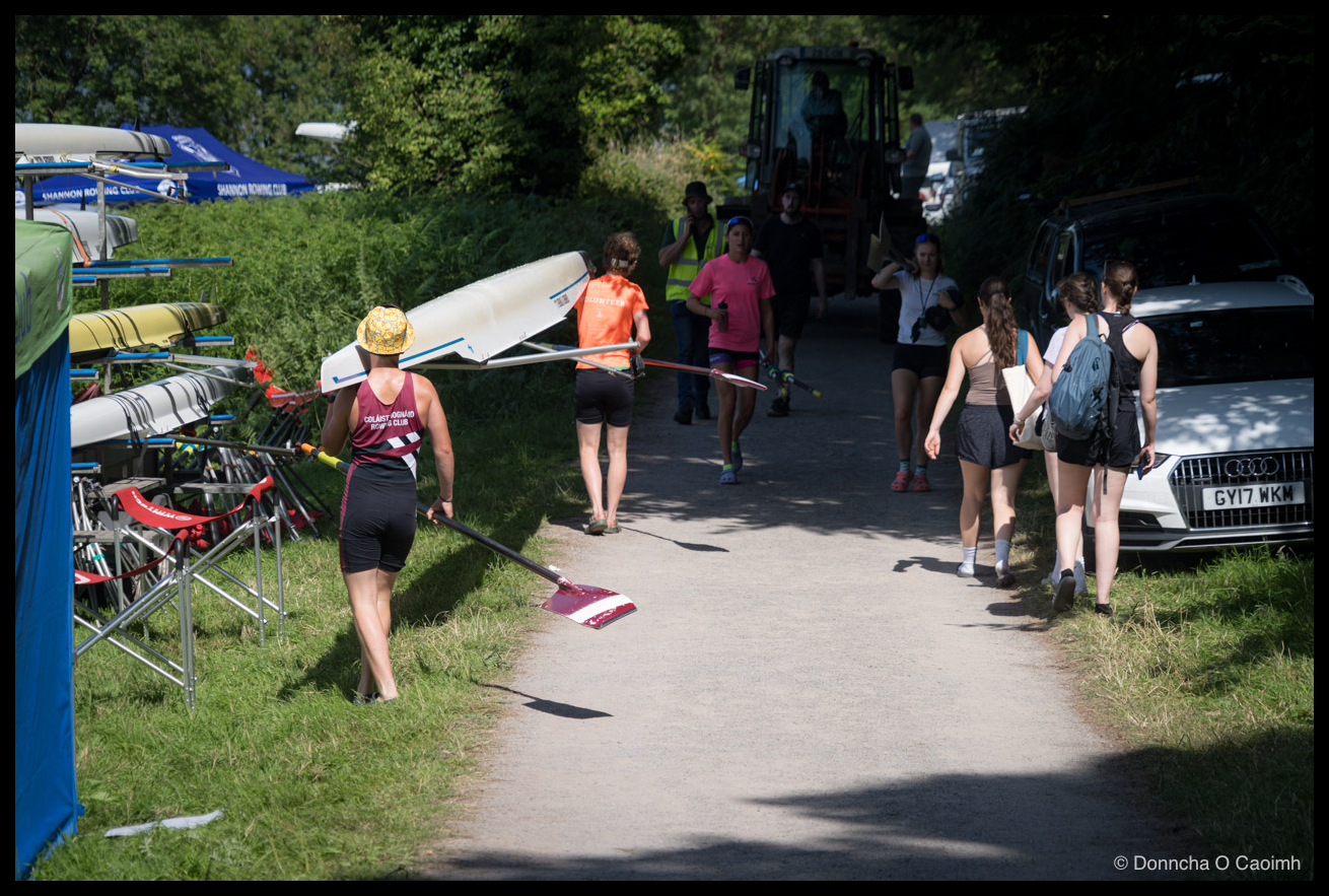 Rowers and volunteers walking along a gravel path at a regatta, a woman in a yellow bucket hat and a maroon "COLÁISTE IOGNÁID ROWING CLUB" singlet carrying an upturned single scull on her shoulder and a blade in her hand, another rower in an orange "VOLUNTEER" shirt carrying blades ahead of her, a blue "SHANNON ROWING CLUB" gazebo and boat racks to the left, and a white Audi with the registration "GY17 WKM" parked on the right beneath a tractor further up the path.