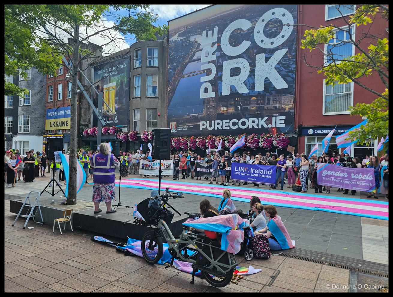 A speaker in a purple "Trans PRIDE CORK" vest addressing a rally from a low stage, with a long trans pride flag rolled out across the pavement and banners reading "LINC Ireland — LBQ Women T&NB Inclusive", "gender rebels — Trans Peer-Support Network" and "WE'RE WITH YOU UKRAINE"; a large "PURE CORK — PURECORK.IE" mural fills the wall behind.