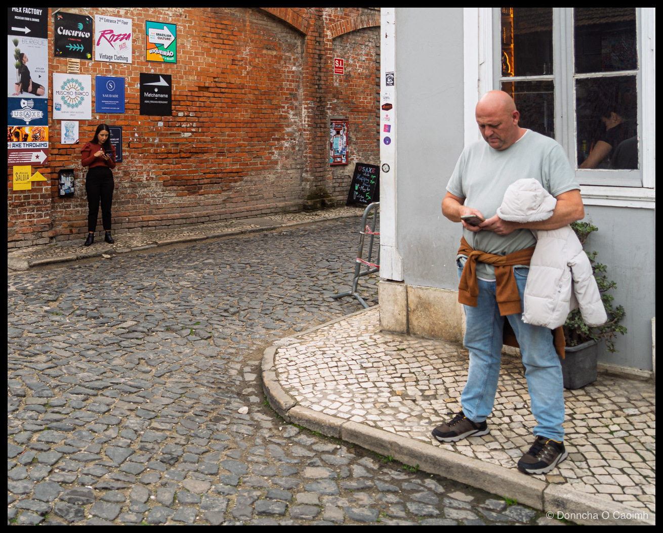 A cobbled street corner in Lisbon where a bald man in a pale green T-shirt, jeans and a tan jumper tied round his waist stands on white calçada paving looking at his phone with a white padded coat over one arm, while across the square a woman in a rust-coloured top and black trousers leans against a red brick wall also looking at her phone, the wall covered in posters reading MEX FACTORY MEXICAN FOOD & DRINKS, BERGUE ATELIER BOUTIQUE, Cucurico, ROZZA SAUCE, Vintage Clothing, 2nd Entrance 2nd Floor, BOQUEIRÃO Brazilian Culture, MUSCHIO BIANCO, SAUDADE, Matchamama, USAXE and AXE THROWING, with a yellow SALOIA arrow sign low on the brick and a red S.I. sign above.