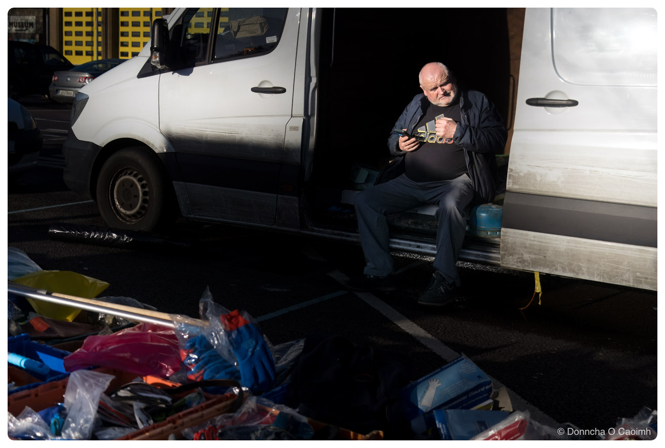 A man sits in the open side door of a white Mercedes Sprinter van in low winter sunlight, wearing a dark jacket over a black Adidas T-shirt and looking down at a phone in his hands, with a stack of stock in plastic bags and boxes of disposable gloves piled on the tarmac in the foreground and a sign reading MUSEUM on the wall behind the van.