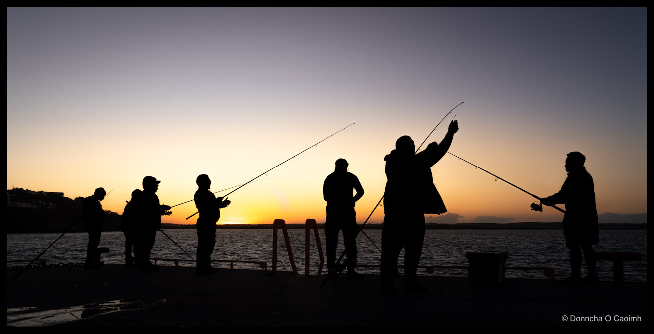 Seven fishermen silhouetted on a wet pier after sunset, with two rods on opposite sides of the frame curving up to form a wide V against an orange and blue sky, a low headland across the water.