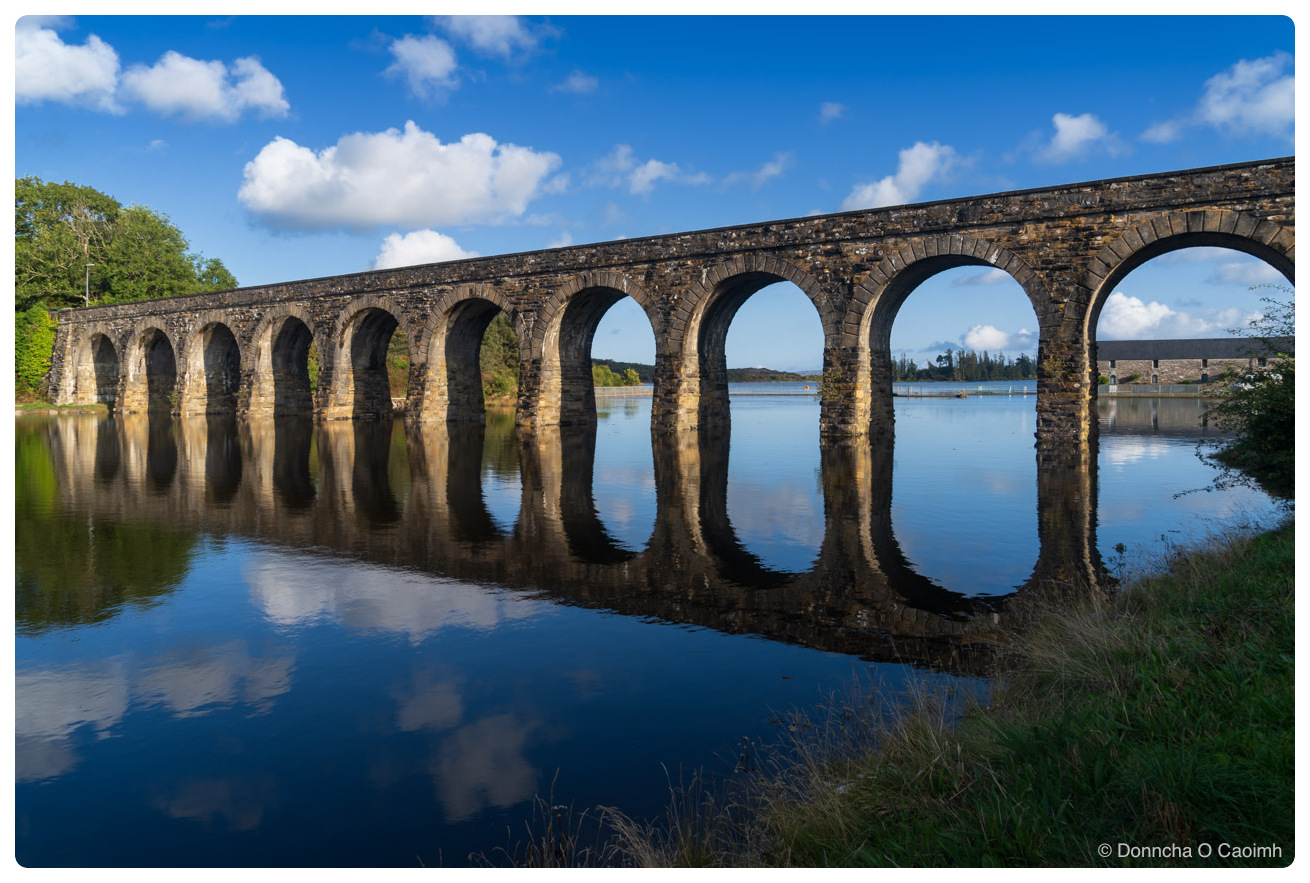 A stone railway viaduct of twelve arches crossing still water on a bright day, its arches and the white cumulus above mirrored almost perfectly in the surface, with trees on the left bank, grass in the foreground and a long grey building visible through the right-hand arches.