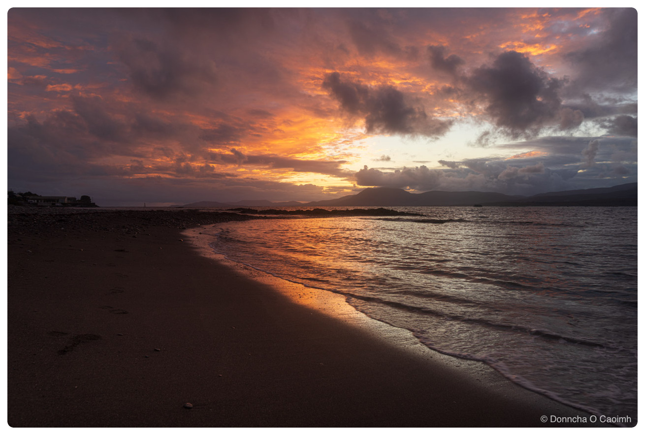 Wet sand and shallow water on a beach at sunset, reflecting a sky of orange and grey cloud, with the dark ridge of a peninsula on the far side of the bay and a few buildings on the shoreline to the left.