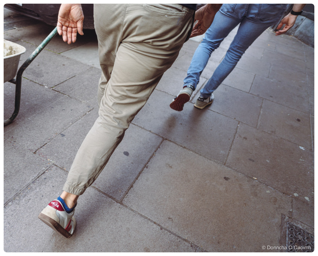 Two people walking away across large grey paving slabs, cropped at the waist, one in beige cuffed trousers and white VEJA runners with red and blue trim, the other in light blue jeans and grey VEJA runners with a red rubber sole lifted mid-step, a green metal barrow leg at the left edge and a cast iron drain cover bottom right.