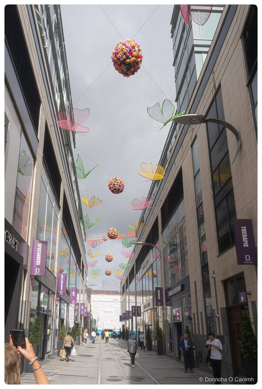 Looking up a narrow pedestrian shopping street with rows of hanging decorations strung on wires between the buildings: large spherical balls of pink, yellow and orange flowers, and wire-frame butterflies and flowers in green, pink and yellow. The sky above is overcast grey with an orange construction crane visible in the distance. Purple banners on the buildings read "SPECSAVERS", "THERAPIE", "SKECHERS" and "OPERA LANE", with shopfront signs for "NEW LOOK" and "Hegarty". Shoppers walk along the paved street below and a woman in the foreground holds up a phone to take a picture.