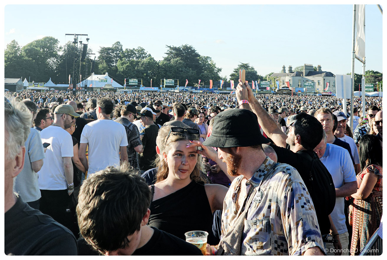 A large festival crowd in a park on a bright summer evening, with people in the foreground queuing for drinks and facing away from the stage, including a bearded man in a black bucket hat and a patterned shirt turning towards a woman in a black one-shoulder top holding a plastic pint of beer, thousands more people stretching back towards the stage rigging and a row of white marquees and signs reading "TOILETS", "FEMALE", "MALE" and "MEDICAL + WELFARE", with mature trees and a large grey house under a clear blue sky behind.